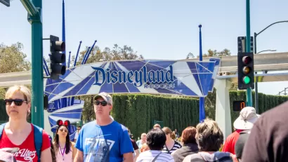 A crowd of people cross the street with a sign for the entrance of Disneyland in the background