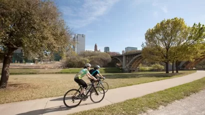 Two cyclists on a paved bike trail through short grass with Dallas buildings in background