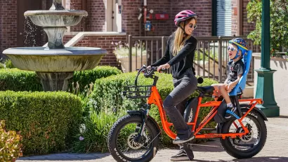 Woman in purple helmet on orange e-bike with baby in seat on back of bike.