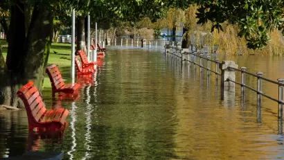 Flood waters overwhelm a park, full of empty benches, on a sunny day.