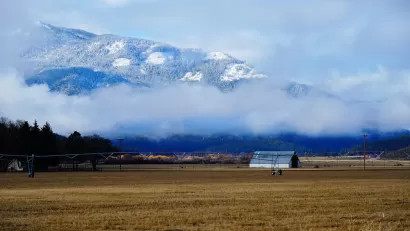 A long farmhouse is visible in winter agricultural fields, Snow-covered mountains loom above low clouds in the background of the photo.