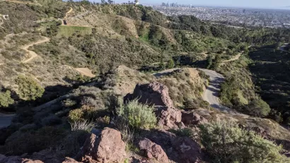 The Griffith Observatory is perched on a hill above Los Angeles, with the city's downtown shown int he distant background. Griffith Park's rolling hills and winding roads are shown in the foreground.