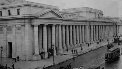 An image of historic Penn Station when it was first built in 1910, viewed from the outside with people, carriages, and streetcars passing by.