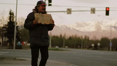 Unhoused man holds up cardboard sign on street corner in Anchorage, Alaska with snowy mountains in background.