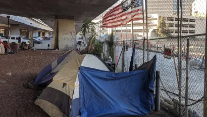 Tents set up by unhoused people under freeway overpass in San Jose, California with American flag above them.