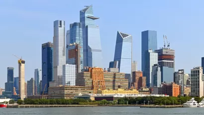 A large group of glass covered skyscrapers rises above the Hudson River in Manhattan, with several new buildings under construction visible too.