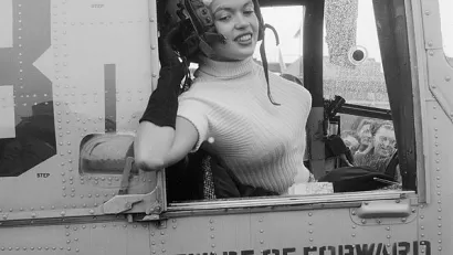 Black and white photo of actress Jayne Mansfield leaning out of the window of a military plane wearing flight helmet for promotional photo