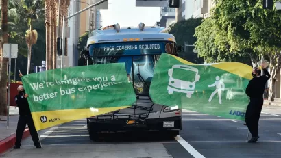 Bus driving through green banner held by two Metro employees at inauguration of new bus lane in downtown Los Angeles.