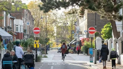 Person on bike on small road in London with people eating at outdoor dining tables and pedestrians walking on either side.