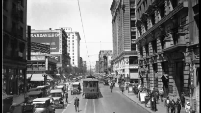 Black-and-white 1924 photo of a street in Los Angeles with a street car, cars driving in the other lane over street car tracks and pedestrians walking on sidewalk and middle of street.