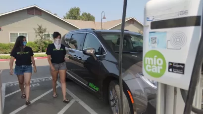 Black SUV charging at Miocar electric car sharing station with two women walking towards it.