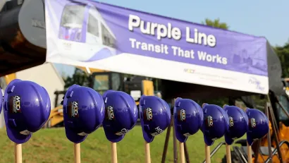 Line of purple construction helmets on shovels in front of Purple Line banner for photo op at BRT line groundbreaking.