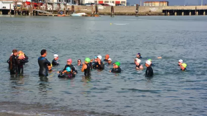 A group of wetsuit-clad swimmers gathers to talk in shallow water near the shore of the San Francisco Bay.