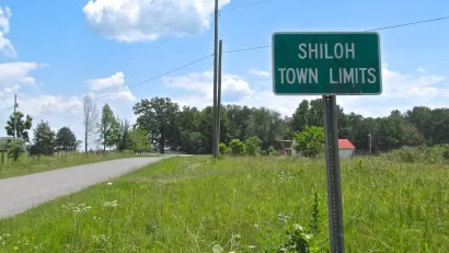 Green sign with white text 'Shiloh Town Limits' against green grassy field with small road on left.