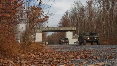 Military humvee driving through gate at Fort Indiantown Gap Natl Guard training center in Pennsylvania surrounded by winter trees and dead leaves.