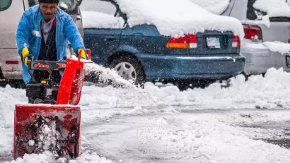 A man pushes a snow blower to clear the ground of an outdoor parking lot filled with cars.