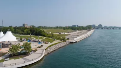 Aerial view of Uniroyal Promenade site on East Riverfront in Detroit with wide paved walkway along river and shaded pavilion at left.