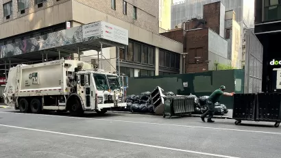 White waste management sanitation garbage truck and worker pushing wheeled green dumpster on street with piles of trash bags.