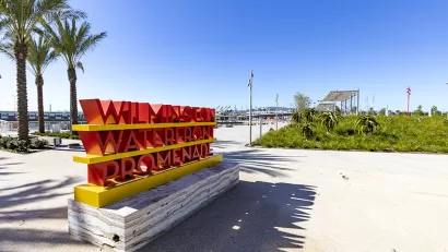 Person-height red 'Wilmington Waterfront Promenade' sign on concrete walkway with palm trees in background on waterfront walkway at the Port of Los Angeles, California.