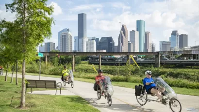 Three men riding recumbent bikes on paved greenway next to bayou with Houston skyline in background.