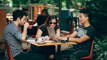 Three young people sitting at a table at outdoor restaurant, one has Apple laptop open.