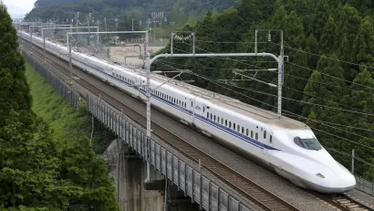 Shinkansen high-speed rail train traveling across bridge with green trees on either side.
