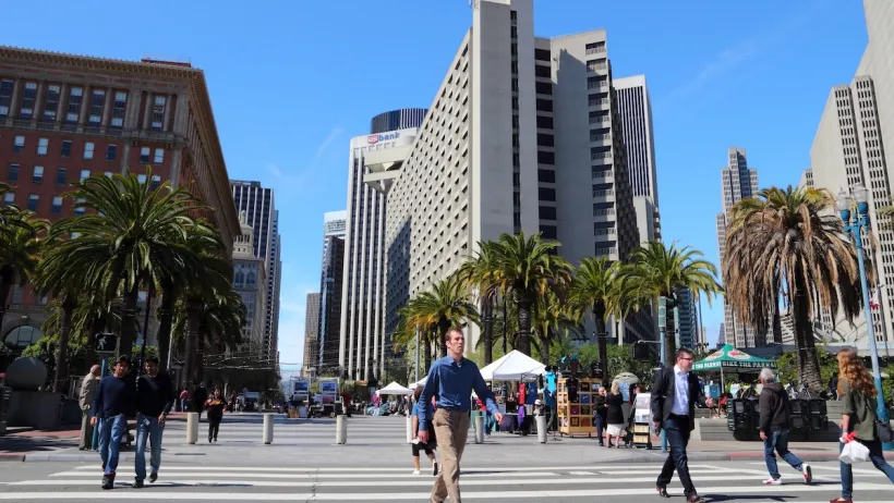 People cross a wide street in San Francisco, with tall buildings in the background.