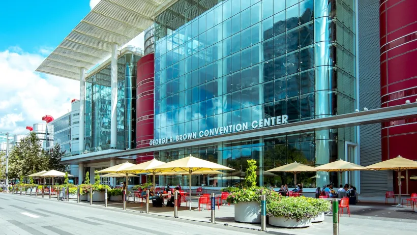 Front of George R. Brown convention center in Houston, Texas with yellow umbrellas over red tables in front.