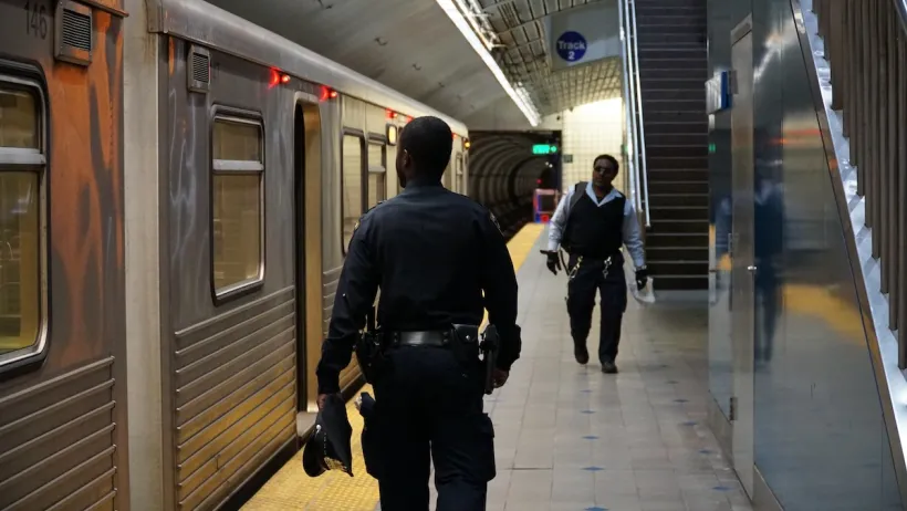 Security officer in dark blue uniform stands next to silver subway train on platform.