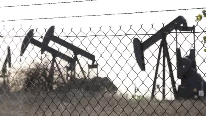 View of black oil wells behind chain link fence with barbed wire top