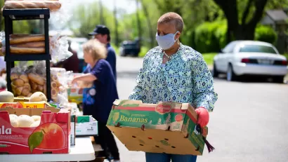 Food bank worker carries box of food in front of outdoor table filled with food items