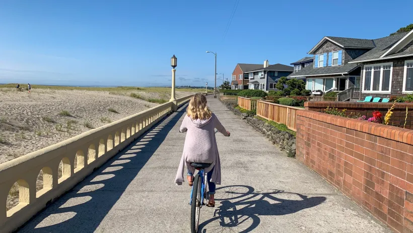 Woman in pink sweater rides a beach cruiser bicycle on a paved trail next to the beach in Seaside, Oregon.