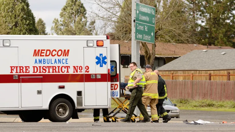 White ambulance and three first responders in yellow vests and firefighting suit wheel gurney into ambulance at car crash site at small intersection.