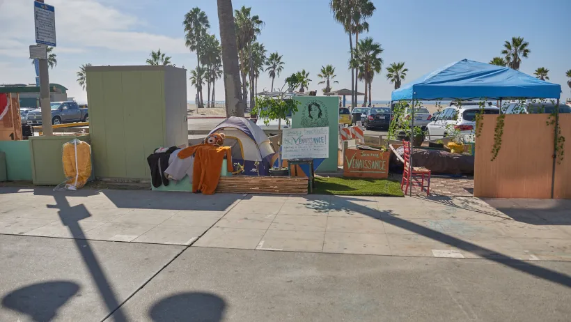 Tents used by unhoused people on concrete walkway with Venice Beach, California beach and palm trees in background.