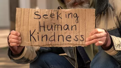 Unhoused man holding sign that reads "Seeking human kindness"