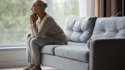 Side view of older woman sitting on gray sofa gazing pensively out of wall-sized window.