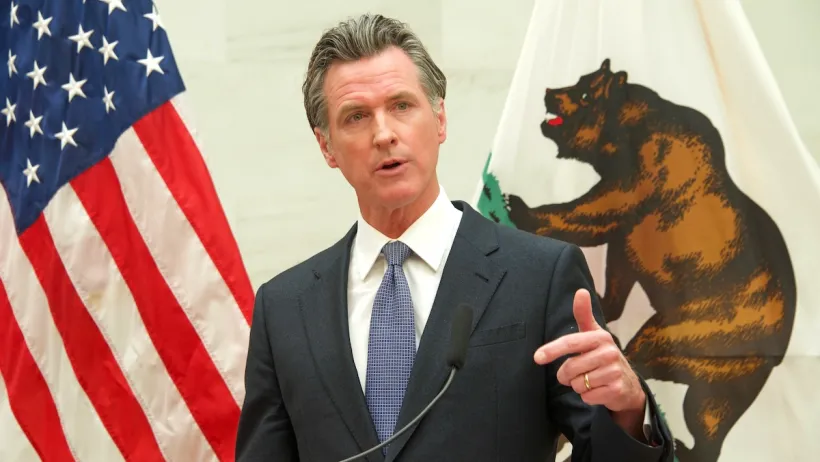 Gavin Newsom standing behind a podium, pointing at the camera. The flag of the United States and the flag of California are behind him.