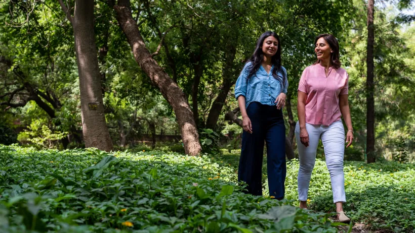 Two women in modern business casual clothing walking through lush green garden or park in Delhi, India.