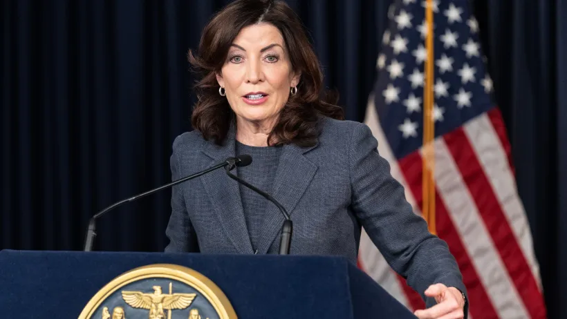 Gov. Kathy Hochul of New York speaking at a podium in front of a dark blue wall and American flag.