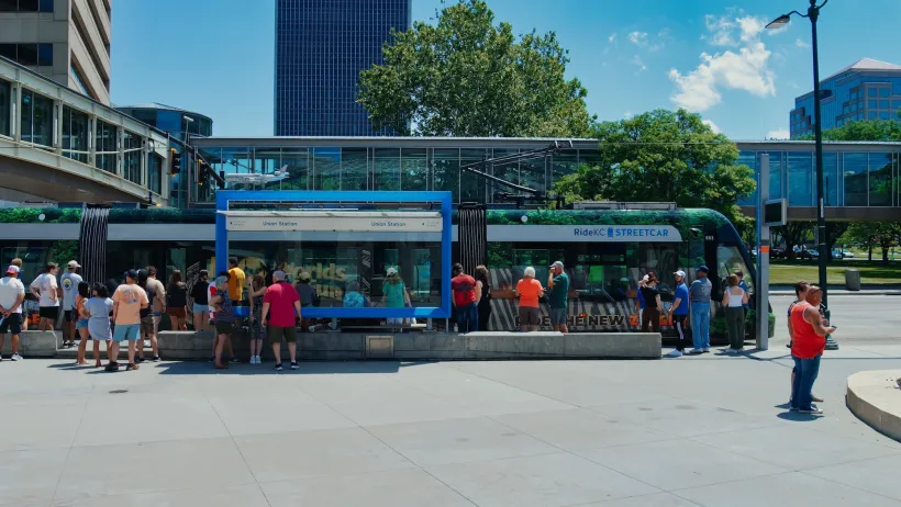 Crowd of people stand at station in front of blue and green Kansas City streetcar.