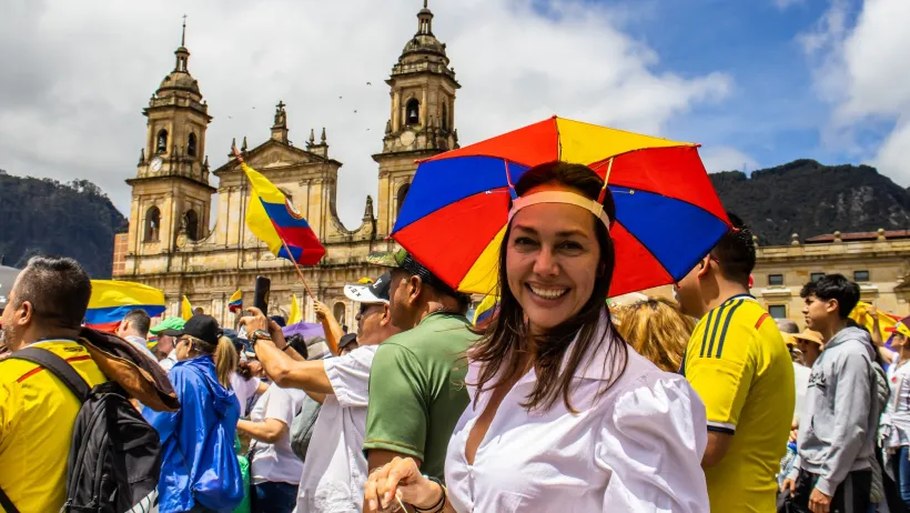 Woman wearing red, yellow, and blue umbrella hat smiling at camera during political protest in front of old Spanish-style yellow church in Bogota, Colombia.
