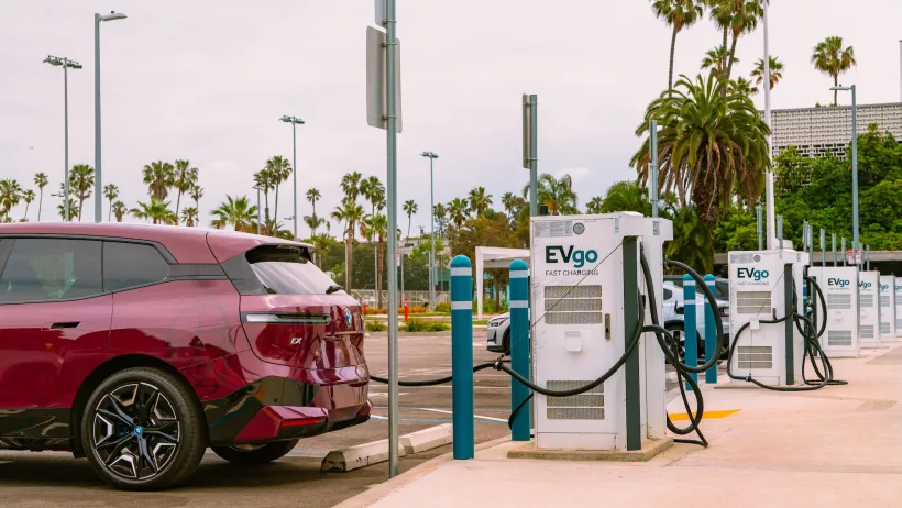 Red electric car plugged into charging station in outdoor parking lot with palm trees in background.