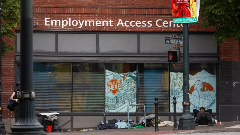 Unhoused people camped in front of brick building with "Employment Access Center" sign in Portland, Oregon.