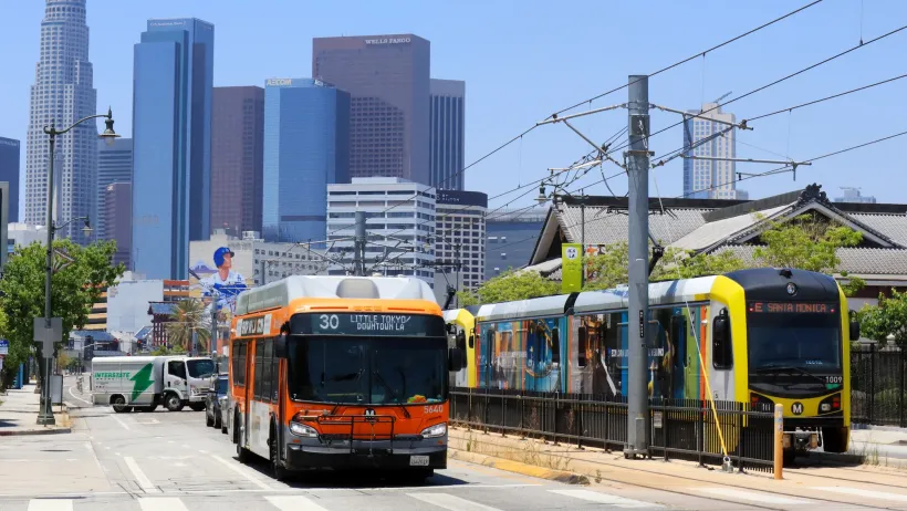 Orange bus and yellow light rail train next to each other on street with downtown Los Angeles visible in background.
