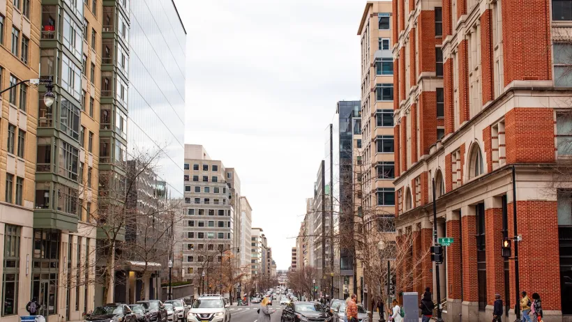 Street scene in Washington DC with mixed modern and brick buildings and pedestrians crossing a crosswalk.