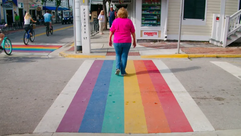 Older white woman in pink shirt and blue jeans walking across Rainbow crosswalk in Key West, Florida.