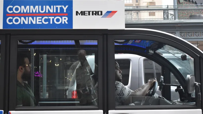 A modern Metro Community Connector (Cab) EV Vehicle, cruising during midday with a passenger in the downtown financial district area.