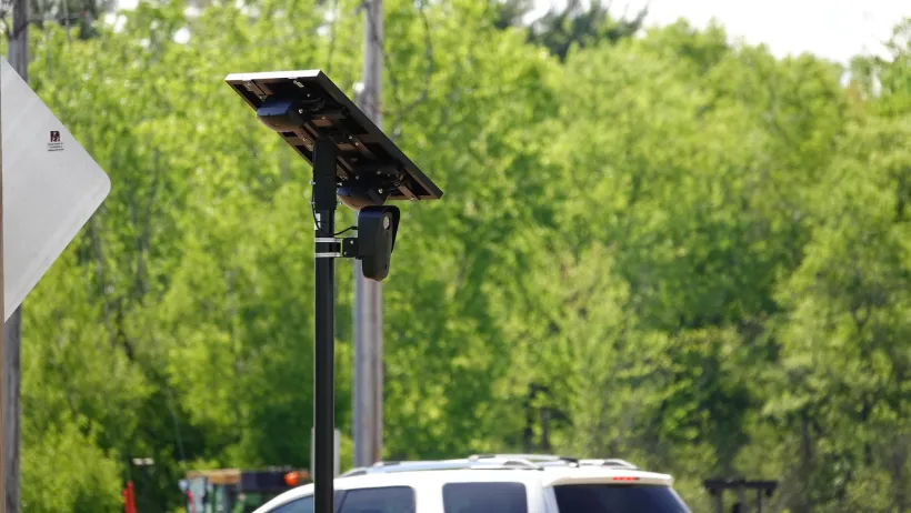 A black Flock traffic camera with solar panel above it installed on a pole outdoors with car visible in background.