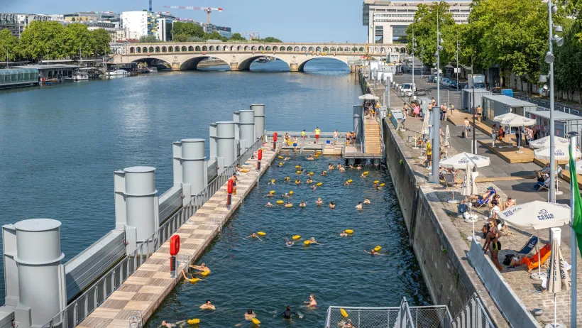 Swimmers in an enclosed swimming area on the Seine River in Paris, France with a low arched bridge visible in background.