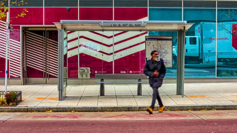 Man sitting at bus stop in Washington DC with red building wall in background.